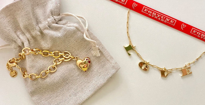 Jeweled Heart Bracelet laying on a bag and a LOVE Letters Necklace next to a Fortune & Frame Ribbon.